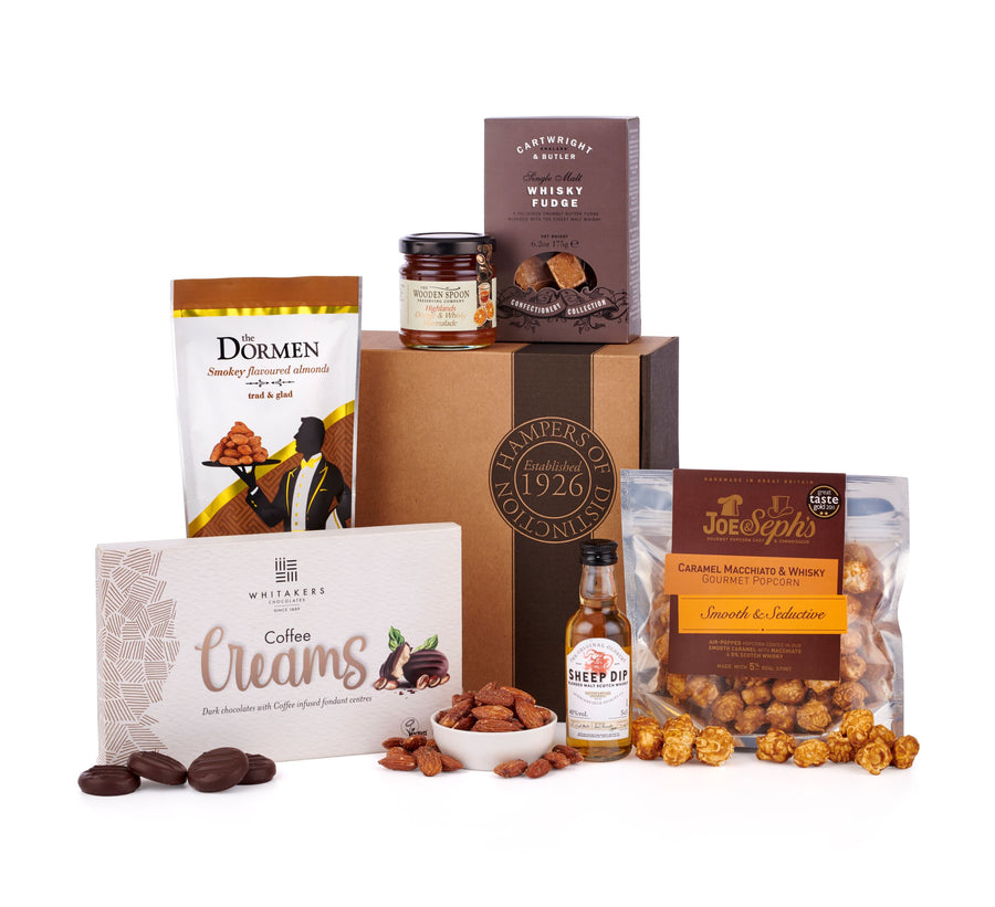 The classic Whisky Lover Gift Box sat on top of a white table top. Centred around the Sheep Dip whisky pictured in the middle of the image, this is surrounded by Coffee Creams, Whisky Fudge by Cartwright & Butler, Almonds, Macchiato & Whisky popcorn & more. This Whisky Hamper & Gift Idea is hand-packed into the brown gift box pictured in the background of the image. 